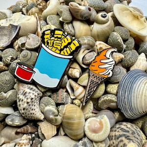 Op de afbeelding: Twee emaillepins die een blauwe en witte container met gele frietjes en een oranje ijshoorn op een bed van schelpen voorstellen.