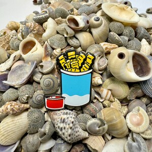 Op de afbeelding: Een gele en blauwe emaille pin van een blikje friet met een rode dipsaus. De pin is omringd door een stapel schelpen.