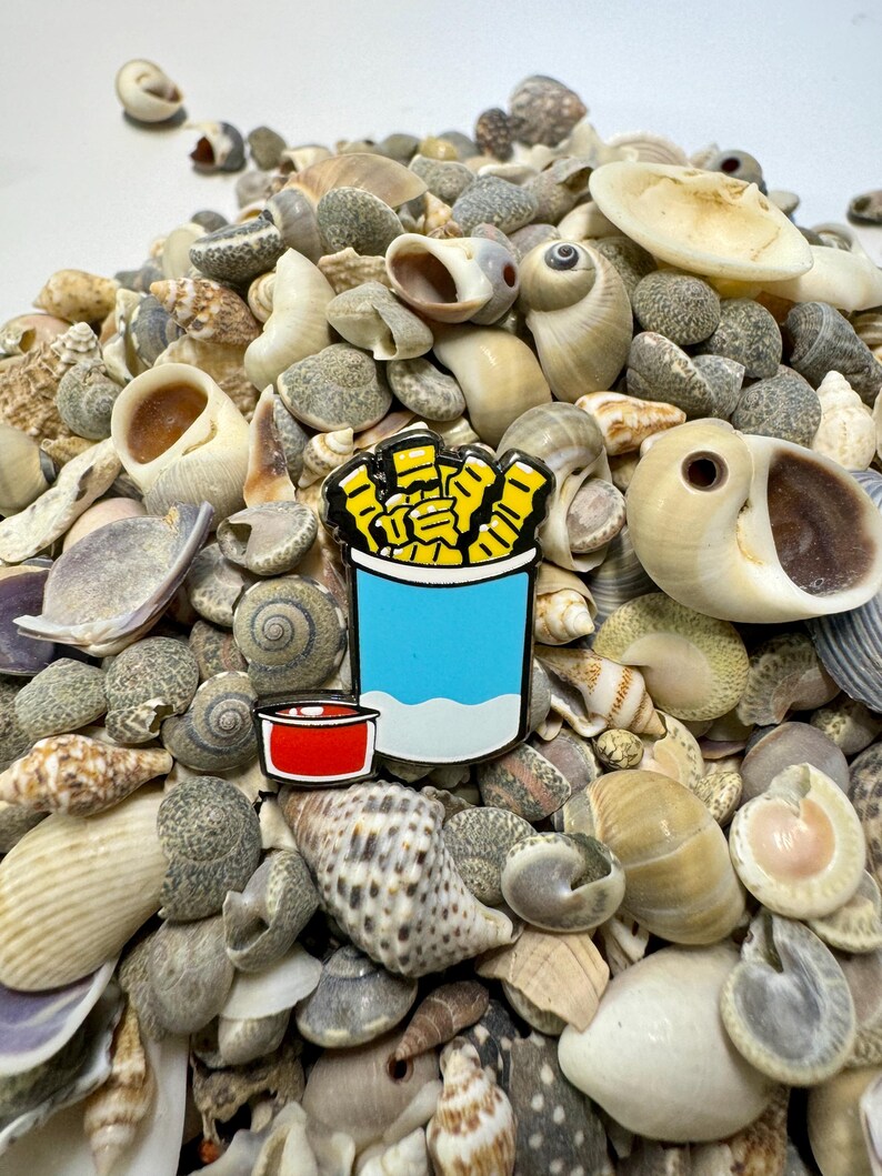 Op de afbeelding: Een gele en blauwe emaille pin van een blikje friet met een rode dipsaus. De pin is omringd door een stapel schelpen.