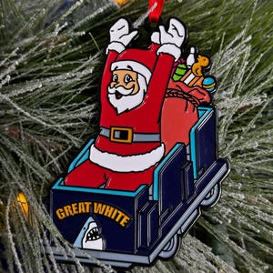 May include: A red and white Santa Claus ornament riding a blue and white roller coaster with the words "Great White" on the side. The roller coaster has a shark fin on the back.