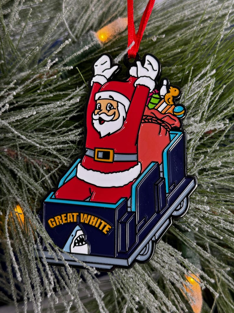 May include: A red and white Santa Claus ornament riding a blue and white roller coaster with the words "Great White" on the side. The roller coaster has a shark fin on the back.