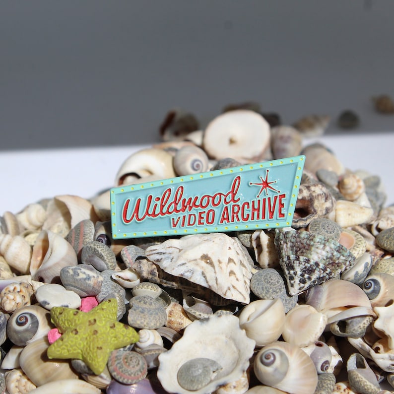 May include: A blue and red enamel pin with the text "Wildwood Video Archive" and a star design. The pin is surrounded by a variety of seashells.