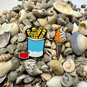 Op de afbeelding: Twee emaillepins die een blauwe en witte container met frietjes met een rood ketchupbekertje en een ijshoorn met een swirl van witte frosting voorstellen. De pins liggen op een stapel schelpen.