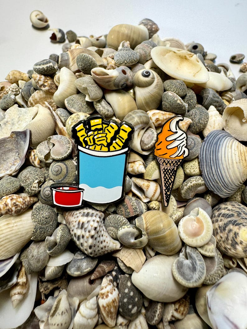 Op de afbeelding: Twee emaillepins die een blauwe en witte container met frietjes met een rood ketchupbekertje en een ijshoorn met een swirl van witte frosting voorstellen. De pins liggen op een stapel schelpen.