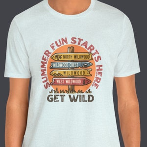 Pu&ograve; includere: Una maglietta grigio chiaro con una grafica circolare che recita "SUMMER FUN STARTS HERE" attorno a un design di palo segnaletico. Il palo segnaletico indica "NORTH WILDWOOD", "WILDWOOD CREST", "WILDWOOD" e "WEST WILDWOOD". Il testo "GET WILD" &egrave; sotto.