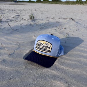 May include: A light blue baseball cap with a dark navy blue brim and a yellow rope detail. The front features a rectangular patch with the text Wildwood New Jersey Boardwalk Beach Summer and a small graphic of a Ferris wheel in the center.