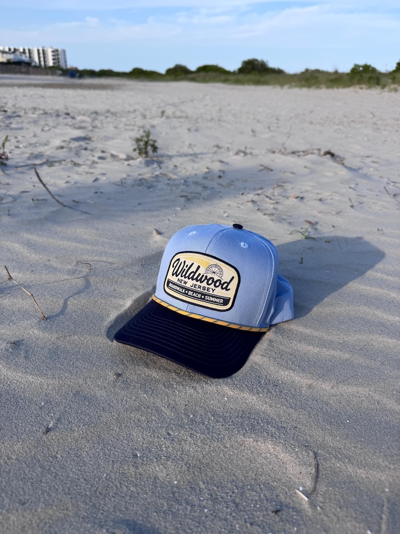 May include: A light blue baseball cap with a dark navy blue brim and a yellow rope detail. The front features a rectangular patch with the text Wildwood New Jersey Boardwalk Beach Summer and a small graphic of a Ferris wheel in the center.