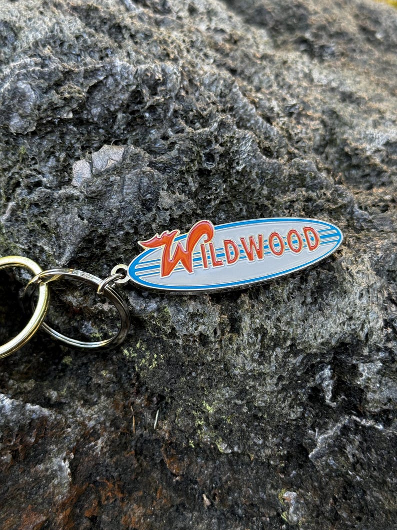 May include: A silver keychain with a blue and white oval charm that says "WILDWOOD" in red letters. The charm has a red and orange wave design.