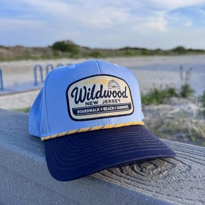 May include: A light blue baseball cap with a dark navy blue brim and a yellow rope detail. The front features a rectangular patch with the text Wildwood New Jersey Boardwalk Beach Summer and a small graphic of a Ferris wheel in black and yellow.