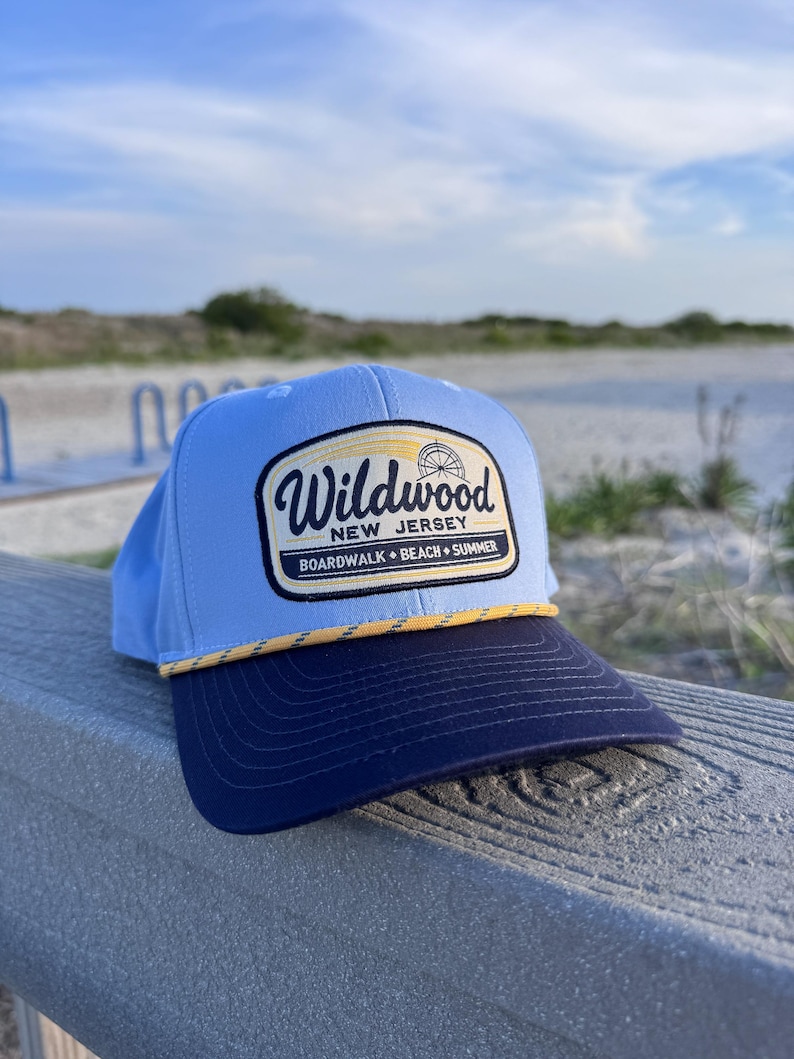 May include: A light blue baseball cap with a dark navy blue brim and a yellow rope detail. The front features a rectangular patch with the text Wildwood New Jersey Boardwalk Beach Summer and a small graphic of a Ferris wheel in black and yellow.