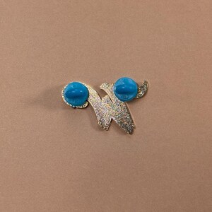 May include: A gold-colored pin with a glittery finish in a stylized letter "W" shape. The pin has two bright turquoise-colored pin backs. The pin is set against a solid brown background.