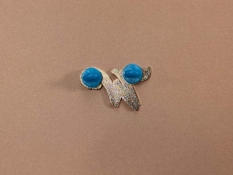 May include: A gold-colored pin with a glittery finish in a stylized letter "W" shape. The pin has two bright turquoise-colored pin backs. The pin is set against a solid brown background.