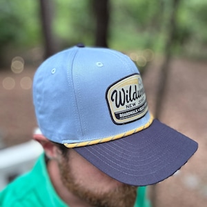 May include: A light blue baseball cap with a dark navy blue brim and a yellow and white braided rope detail. The front features a rectangular patch with the text Wildwood New Jersey Boardwalk Beach in black and yellow lettering on a white background.
