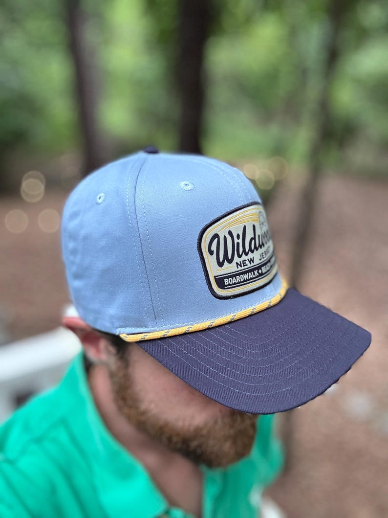 May include: A light blue baseball cap with a dark navy blue brim and a yellow and white braided rope detail. The front features a rectangular patch with the text Wildwood New Jersey Boardwalk Beach in black and yellow lettering on a white background.
