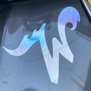 May include: A silver iridescent sticker with the letter "W" in a stylized font. The sticker is on a gray car.