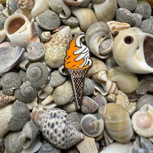 Op de afbeelding: Oranje en witte emaille pin van een ijshoorn op een bed van schelpen.