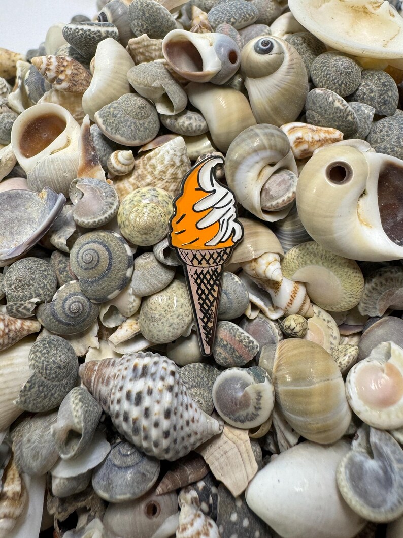 Op de afbeelding: Oranje en witte emaille pin van een ijshoorn op een bed van schelpen.