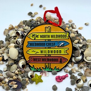 May include: A yellow, round, metal sign with four directional arrows pointing to different locations. The arrows are labeled "North Wildwood", "Wildwood Crest", "Wildwood", and "West Wildwood". The sign is surrounded by seashells.