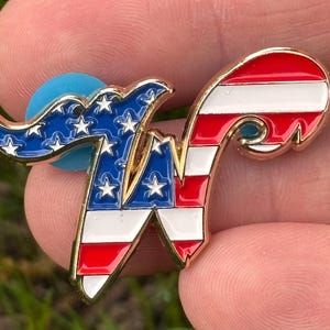 May include: A gold-toned metal pin in the shape of the letter "W", featuring a patriotic design. The left side displays a blue field with white stars, while the right side has red and white stripes. A blue backing is visible.
