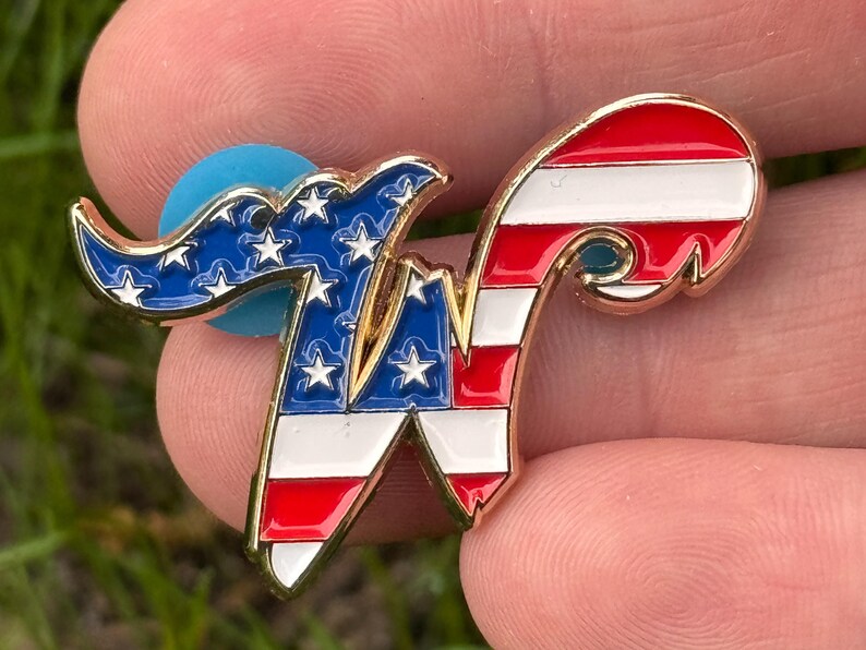May include: A gold-toned metal pin in the shape of the letter "W", featuring a patriotic design. The left side displays a blue field with white stars, while the right side has red and white stripes. A blue backing is visible.