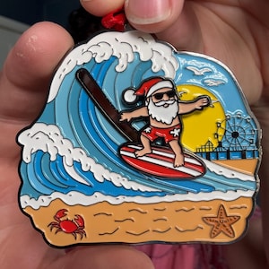 Peut inclure: Un ornement décoratif en métal représentant un père Noël surfeur portant des lunettes de soleil et un short de bain rouge sur une planche de surf rouge et blanche. La scène inclut une grande vague bleue, un soleil jaune, une jetée avec une grande roue, un crabe rouge et une étoile de mer.