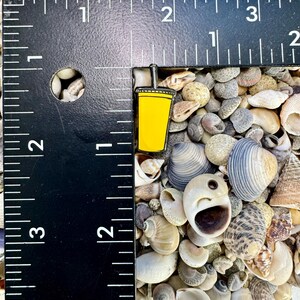 K&ouml;nnte beinhalten: Eine gelbe Emaille-Anstecknadel mit einem Becher mit Strohhalm. Die Anstecknadel ist etwa 2,5 cm hoch und liegt auf einem Hintergrund aus Muscheln.