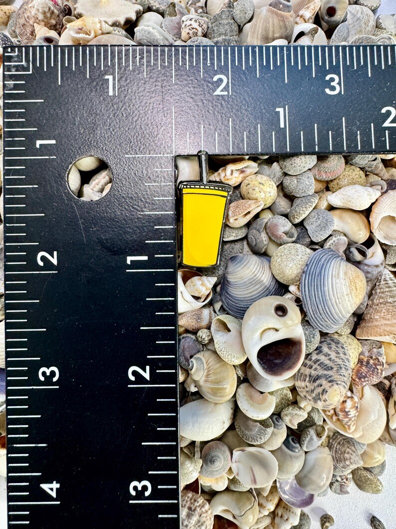 K&ouml;nnte beinhalten: Eine gelbe Emaille-Anstecknadel mit einem Becher mit Strohhalm. Die Anstecknadel ist etwa 2,5 cm hoch und liegt auf einem Hintergrund aus Muscheln.