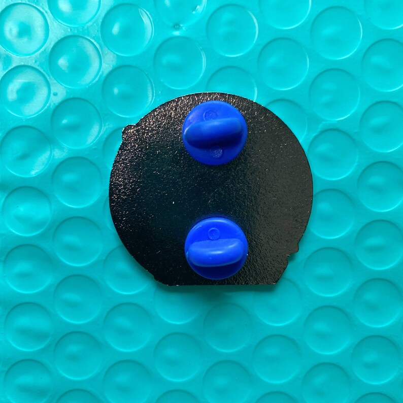 Pu&ograve; includere: Spilla in smalto nero con due perni in plastica blu sul retro.