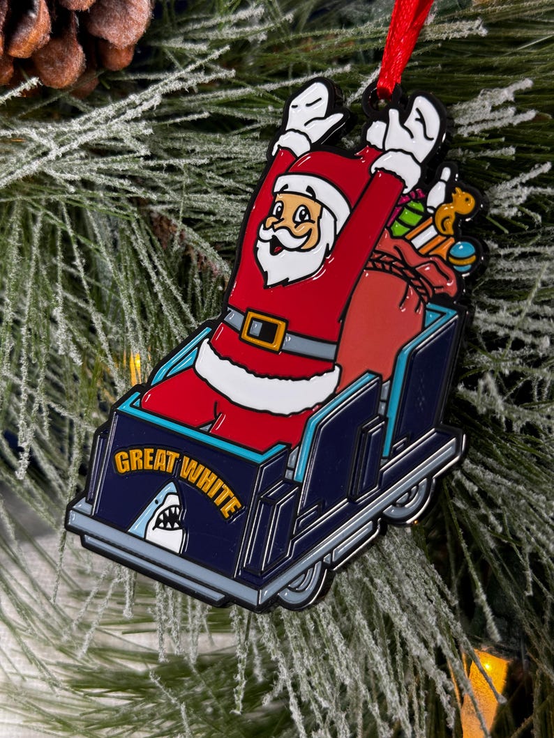 May include: A red and white Santa Claus ornament riding a blue and white roller coaster with the words "Great White" on the side. The ornament is hanging from a red ribbon.