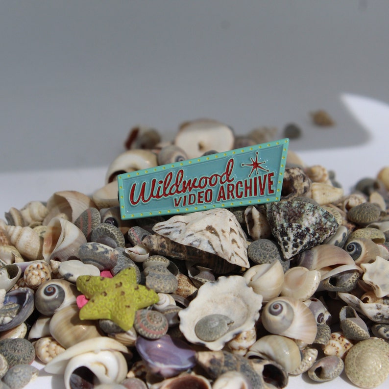 May include: A teal blue enamel pin with the text "Wildwood Video Archive" in red and white lettering. The pin features a starburst design and is surrounded by a variety of seashells.