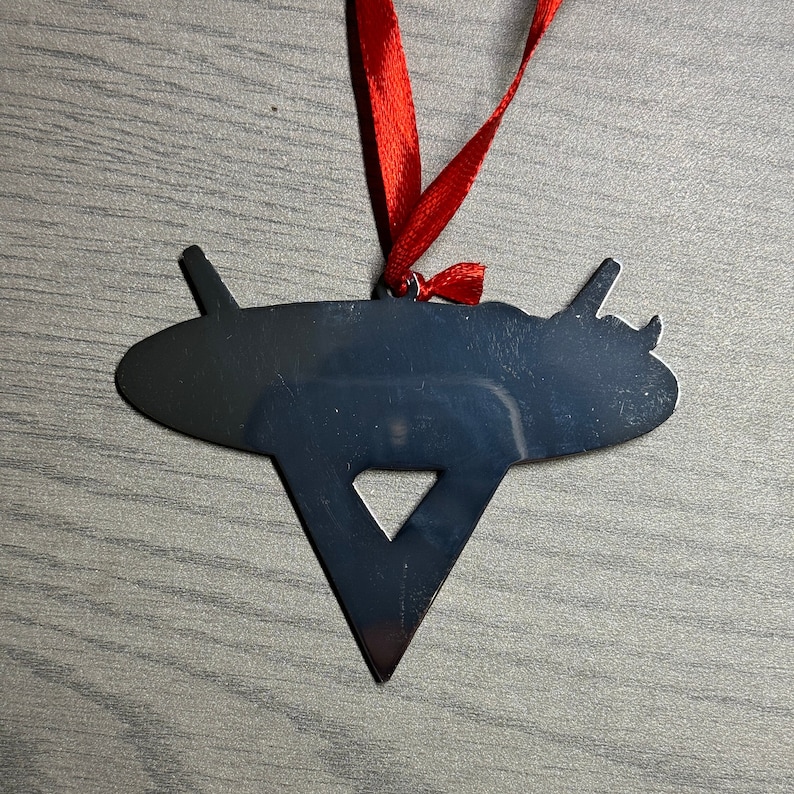 May include: A black metal ornament shaped like a flying saucer with a triangular base. The ornament is attached to a red ribbon.