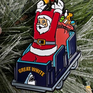 May include: A blue and white Christmas ornament featuring Santa Claus riding in a sleigh with a shark fin and the text "Great White".