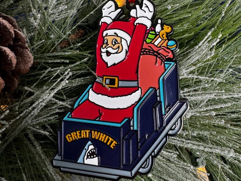 May include: A blue and white Christmas ornament featuring Santa Claus riding in a sleigh with a shark fin and the text "Great White".