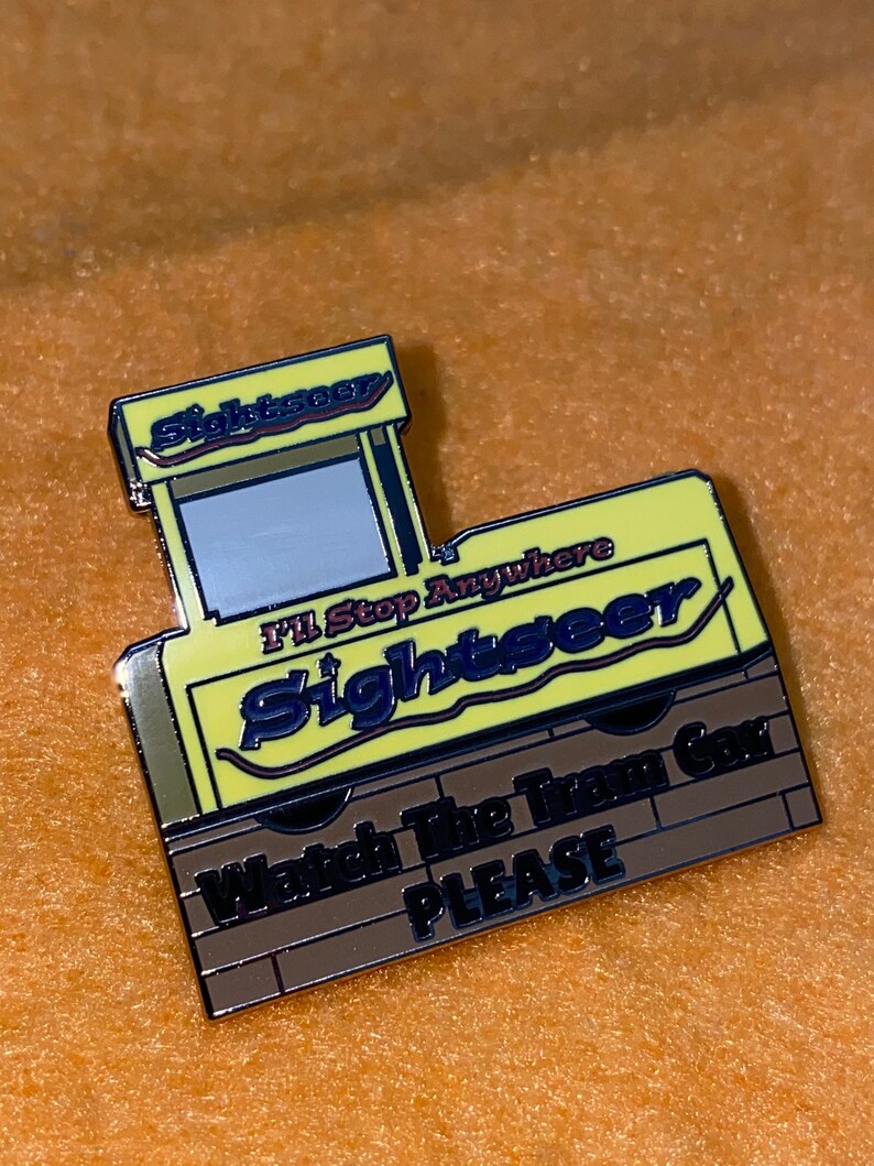 Puede incluir: Un pin de esmalte amarillo y marr&oacute;n con forma de tranv&iacute;a tur&iacute;stico. El pin tiene la palabra "Sightseer" en azul y el texto "I'll Stop Anywhere" y "Watch The Tram Car PLEASE".