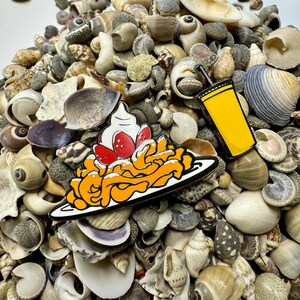 K&ouml;nnte beinhalten: Zwei Emaille-Pins auf einem Haufen von Muscheln. Eine ist eine gelbe Tasse mit einem Strohhalm und die andere ist ein Teller mit orangefarbenen Spaghetti mit roten Erdbeeren und wei&szlig;er Schlagsahne.