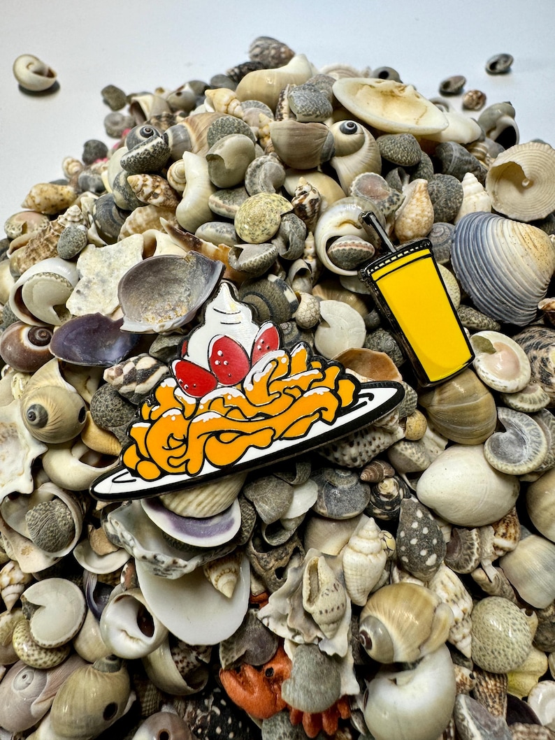 K&ouml;nnte beinhalten: Zwei Emaille-Pins auf einem Haufen von Muscheln. Eine ist eine gelbe Tasse mit einem Strohhalm und die andere ist ein Teller mit orangefarbenen Spaghetti mit roten Erdbeeren und wei&szlig;er Schlagsahne.