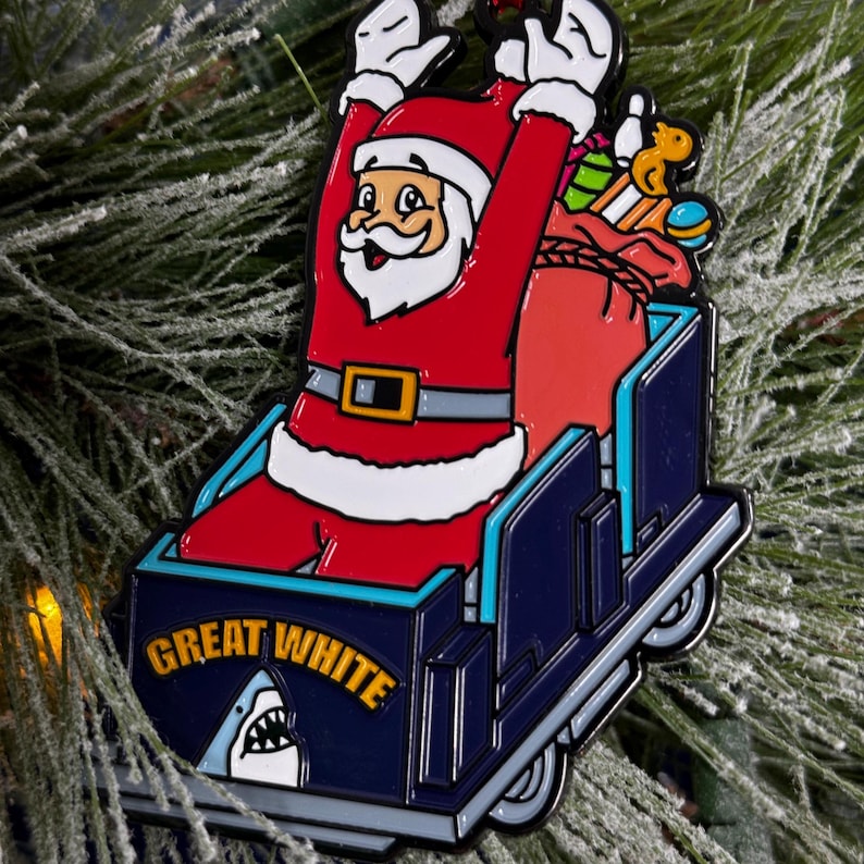 May include: A Christmas ornament featuring Santa Claus riding a blue and white sled with the words "Great White" and a shark fin. Santa is wearing a red suit with a white trim and a black belt. He is holding a bag of gifts and there are toys in the sled.
