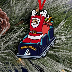 May include: A red and white Santa Claus ornament riding a blue and white sled with a shark fin and the words "Great White" on the side. The ornament is hanging on a green Christmas tree.