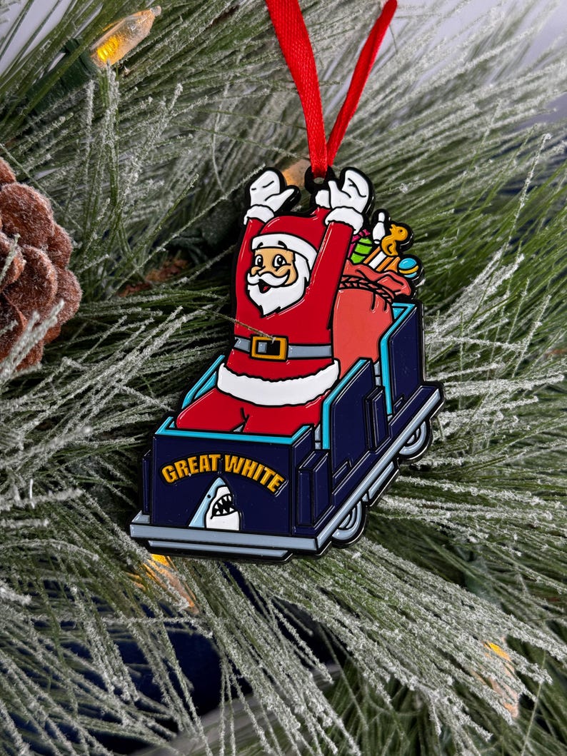 May include: A red and white Santa Claus ornament riding a blue and white sled with a shark fin and the words "Great White" on the side. The ornament is hanging on a green Christmas tree.
