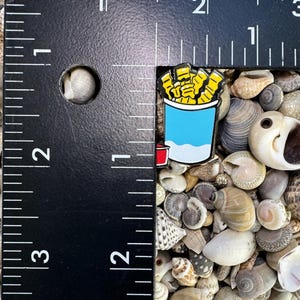 Op de afbeelding: Een close-up van een kleine emaille pin met een cartoonillustratie van een blauwe en witte container met friet. De pin ligt op een bedje van kleine schelpen.