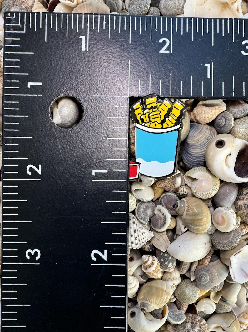 Op de afbeelding: Een close-up van een kleine emaille pin met een cartoonillustratie van een blauwe en witte container met friet. De pin ligt op een bedje van kleine schelpen.