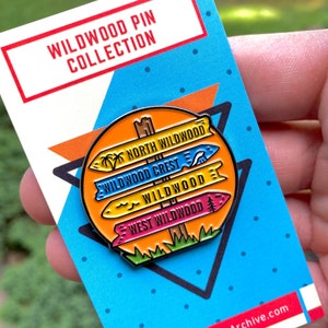 Pu&ograve; includere: Una spilla smaltata colorata con un cartello stradale con quattro frecce direzionali che puntano verso diverse aree di Wildwood, New Jersey. Le frecce indicano "North Wildwood", "Wildwood Crest", "Wildwood" e "West Wildwood".