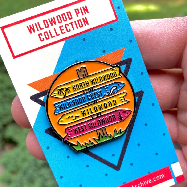 Pu&ograve; includere: Una spilla smaltata colorata con un cartello stradale con quattro frecce direzionali che puntano verso diverse aree di Wildwood, New Jersey. Le frecce indicano "North Wildwood", "Wildwood Crest", "Wildwood" e "West Wildwood".