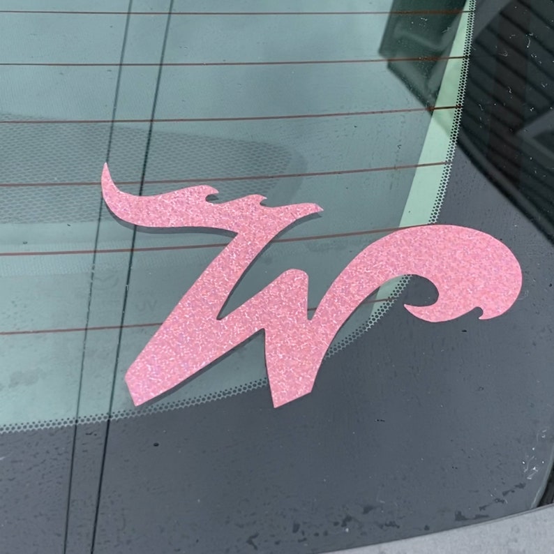 May include: Pink glitter vinyl decal of the letter W with a decorative flourish. The decal is on a car window.