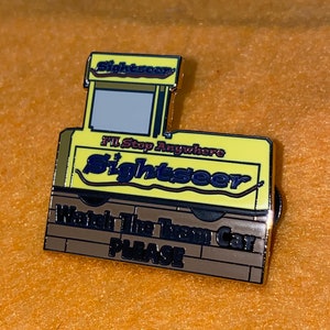 Puede incluir: Un pin de esmalte amarillo y marr&oacute;n con forma de tranv&iacute;a. El pin presenta la palabra "Sightseer" y la frase "I'll Stop Anywhere". La parte inferior del pin dice "Watch The Tram Car PLEASE".
