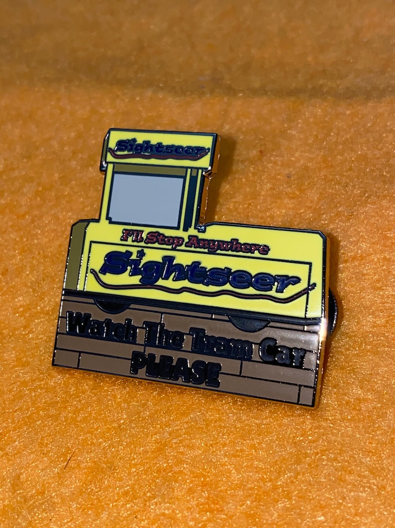 Puede incluir: Un pin de esmalte amarillo y marr&oacute;n con forma de tranv&iacute;a. El pin presenta la palabra "Sightseer" y la frase "I'll Stop Anywhere". La parte inferior del pin dice "Watch The Tram Car PLEASE".