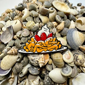 K&ouml;nnte beinhalten: Eine farbige Emaille-Anstecknadel, die einen Teller Pasta mit einem Klecks Schlagsahne und Erdbeeren darauf zeigt. Die Anstecknadel ist von einem Haufen Muscheln umgeben.