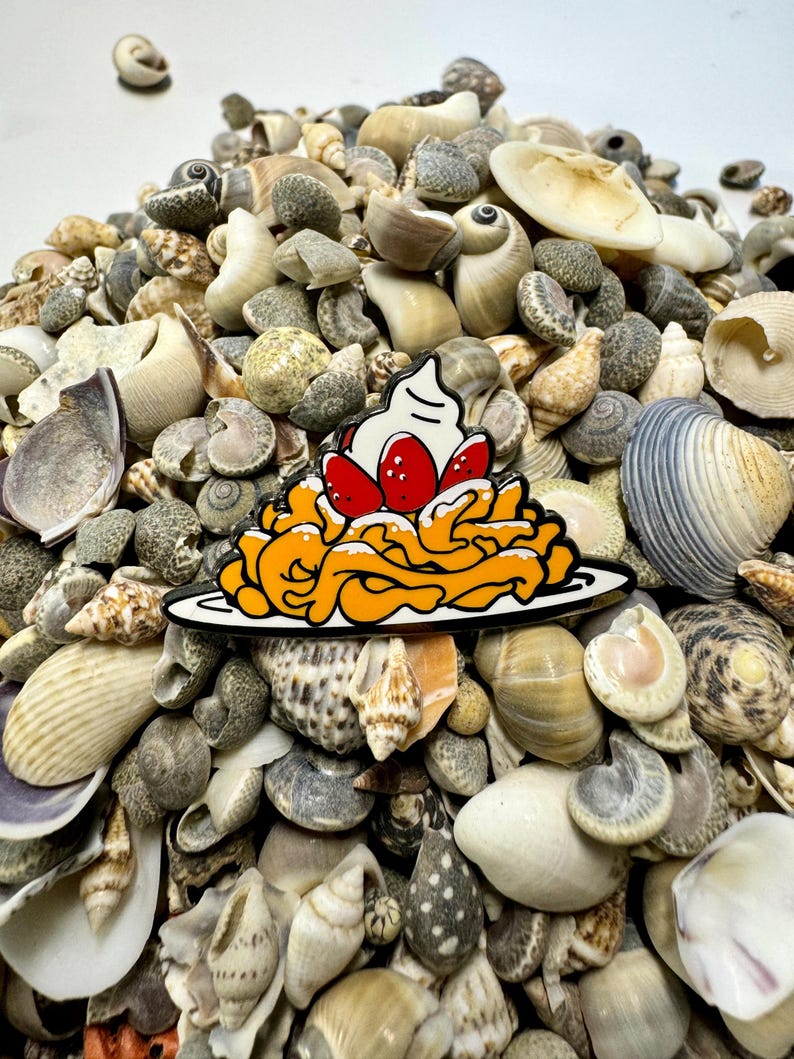 K&ouml;nnte beinhalten: Eine farbige Emaille-Anstecknadel, die einen Teller Pasta mit einem Klecks Schlagsahne und Erdbeeren darauf zeigt. Die Anstecknadel ist von einem Haufen Muscheln umgeben.
