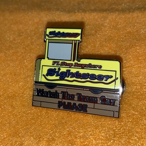 Puede incluir: Un pin de esmalte amarillo y marr&oacute;n con forma de tranv&iacute;a. El pin tiene las palabras "Sightseer" y "I'll Stop Anywhere". La parte inferior del pin dice "Watch The Tram Car PLEASE".