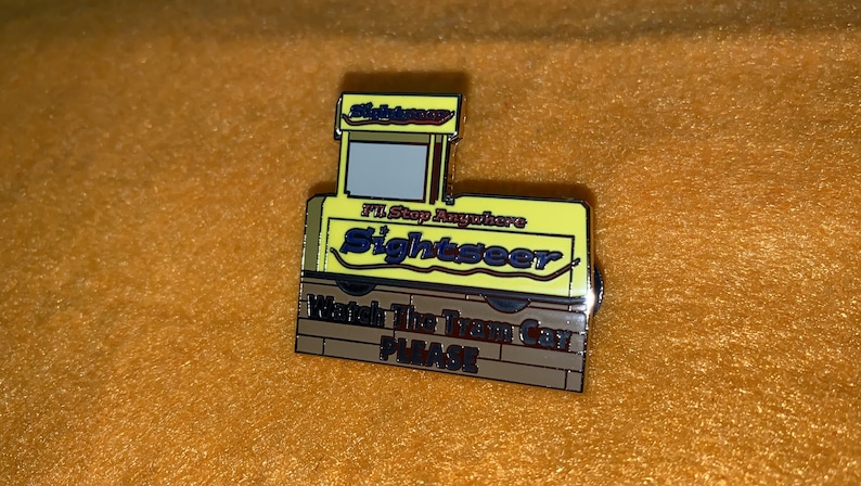 Puede incluir: Un pin de esmalte amarillo y marr&oacute;n con forma de tranv&iacute;a. El pin tiene las palabras "Sightseer" y "I'll Stop Anywhere". La parte inferior del pin dice "Watch The Tram Car PLEASE".